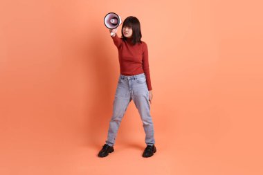 Kırmızı uzun kollu tişörtlü kısa saçlı Asyalı kadın. Bağırma hareketi yapıyor. Elinde turuncu arka planda izole edilmiş bir megafon tutuyor. Gen Z