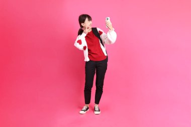 Kırmızı tişörtlü Asyalı kız öğrenci ya da günlük kıyafetler, kırmızı kalp şeklinde gömlek okul çantası taşıyor ve pembe arka planda Holding cep telefonu var. Gen Z