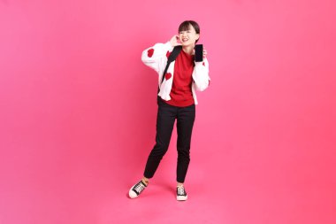 Kırmızı tişörtlü Asyalı kız öğrenci ya da günlük kıyafetler, kırmızı kalp şeklinde gömlek okul çantası taşıyor ve pembe arka planda Holding cep telefonu var. Gen Z