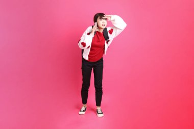 Kırmızı tişörtlü Asyalı kız öğrenci ya da günlük kıyafetler, kırmızı kalp şeklinde gömlek okul çantası taşıyor ve pembe arka planda Holding cep telefonu var. Gen Z