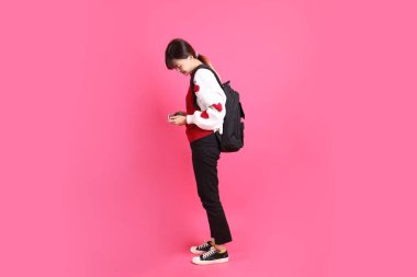 Kırmızı tişörtlü Asyalı kız öğrenci ya da günlük kıyafetler, kırmızı kalp şeklindeki gömlek okul çantası taşıyor ve pembe arka planda el bilgisayarı konsolu oynuyor. Gen Z