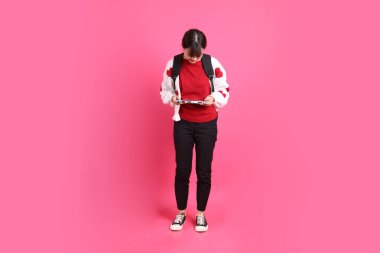 Kırmızı tişörtlü Asyalı kız öğrenci ya da günlük kıyafetler, kırmızı kalp şeklindeki gömlek okul çantası taşıyor ve pembe arka planda el bilgisayarı konsolu oynuyor. Gen Z