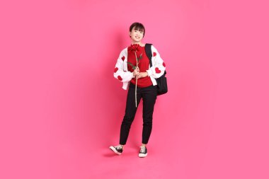 Kırmızı tişörtlü Asyalı kız öğrenci ya da günlük kıyafetler, kırmızı kalp şeklinde gömlek okul çantası taşıyor ve pembe arka planda çiçekleri tutuyor. Gen Z