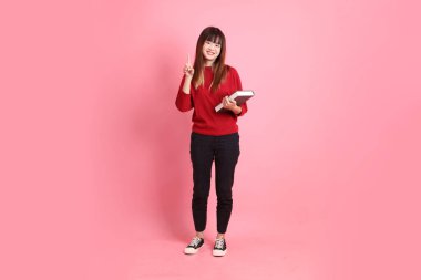 Kırmızı tişörtlü Asyalı kız öğrenci ya da günlük giysiler içinde okul çantası ve pembe arka planda izole edilmiş işaret eden kitaplar taşıyorlar. Gen Z