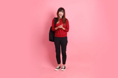 Kırmızı tişörtlü ya da sıradan giysili Asyalı kız öğrenci pembe arka planda etkileyici bir şekilde izole edilmiş okul çantası taşıyor. Gen Z