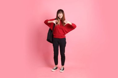 Kırmızı tişörtlü Asyalı kız öğrenci ya da günlük kıyafetler okul çantası ve pembe arka planda izole edilmiş baş parmak hareketleriyle kitap taşıyor. Gen Z