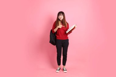 Kırmızı tişörtlü Asyalı kız öğrenci ya da günlük giysiler içinde okul çantası ve pembe arka planda izole edilmiş işaret eden kitaplar taşıyorlar. Gen Z