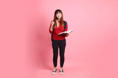 Kırmızı tişörtlü Asyalı kız öğrenci ya da günlük giysiler içinde okul çantası ve pembe arka planda izole edilmiş işaret eden kitaplar taşıyorlar. Gen Z