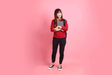 Kırmızı tişörtlü Asyalı kız öğrenci ya da günlük kıyafetler okul çantası ve pembe arka planda sıralanmış kitaplar taşıyor. Gen Z