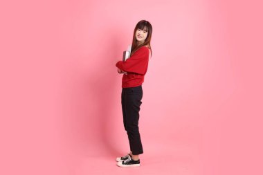 Kırmızı tişörtlü Asyalı kız öğrenci ya da günlük kıyafetler okul çantası ve pembe arka planda sıralanmış kitaplar taşıyor. Gen Z