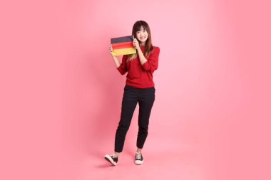 Kırmızı tişörtlü ya da sıradan giysili Asyalı kız öğrenci, pembe arka planda izole edilmiş Alman bayrağı ya da Alman bayrağı taşıyan okul çantası taşıyor. Gen Z