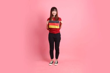 Kırmızı tişörtlü ya da sıradan giysili Asyalı kız öğrenci, pembe arka planda izole edilmiş Alman bayrağı ya da Alman bayrağı taşıyan okul çantası taşıyor. Gen Z