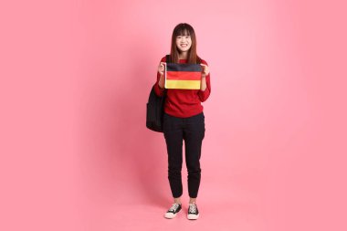 Kırmızı tişörtlü ya da sıradan giysili Asyalı kız öğrenci, pembe arka planda izole edilmiş Alman bayrağı ya da Alman bayrağı taşıyan okul çantası taşıyor. Gen Z