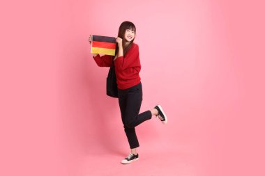 Kırmızı tişörtlü ya da sıradan giysili Asyalı kız öğrenci, pembe arka planda izole edilmiş Alman bayrağı ya da Alman bayrağı taşıyan okul çantası taşıyor. Gen Z