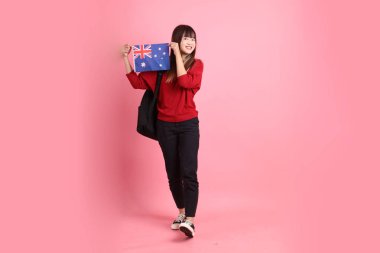 Kırmızı tişörtlü ya da sıradan giysili Asyalı bir kız öğrenci pembe arka planda izole edilmiş Avustralya bayrağı taşıyan bir jestle okul çantası taşıyor. Gen Z