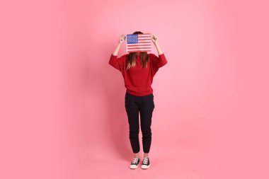 Kırmızı tişörtlü Asyalı kız öğrenci ya da günlük kıyafetler içinde ABD ya da Amerika Birleşik Devletleri ya da pembe arka planda izole edilmiş Amerikan bayrağı taşıyan okul çantası taşıyorlar. Gen Z