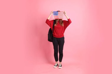 Kırmızı tişörtlü Asyalı kız öğrenci ya da günlük kıyafetler içinde ABD ya da Amerika Birleşik Devletleri ya da pembe arka planda izole edilmiş Amerikan bayrağı taşıyan okul çantası taşıyorlar. Gen Z