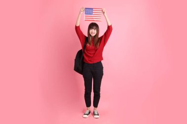 Kırmızı tişörtlü Asyalı kız öğrenci ya da günlük kıyafetler içinde ABD ya da Amerika Birleşik Devletleri ya da pembe arka planda izole edilmiş Amerikan bayrağı taşıyan okul çantası taşıyorlar. Gen Z