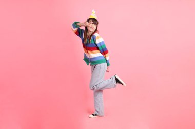 Mutlu yıllar, renkli gökkuşağı kış elbiseli Asyalı kadın, sıradan görünüşlü, barış işareti olan doğum günü şapkası, V işareti, pembe arka planda iki parmak. Gen Z