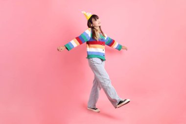 Mutlu yıllar, renkli gökkuşağı kış elbiseli Asyalı kadın, sıradan görünüşlü, pembe arka planda izole edilmiş yürüme jestli doğum günü şapkası. Gen Z, 