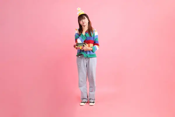 Mutlu yıllar, renkli gökkuşağı kış elbiseli Asyalı kadın, sıradan görünüşlü, doğum günü şapkası ve pembe arka planda saklanmış bir hediye kutusu. Gen Z