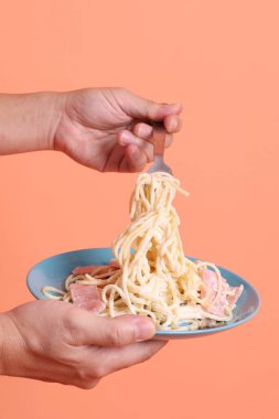 Turuncu arka planda izole edilmiş yeşil bir tabakta, çatallı kremalı spagetti yiyen. İtalyan yemeği, İtalyan mutfağı, erişte..