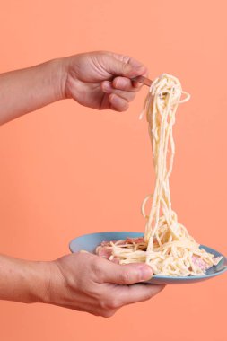 Turuncu arka planda izole edilmiş yeşil bir tabakta, çatallı kremalı spagetti yiyen. İtalyan yemeği, İtalyan mutfağı, erişte..