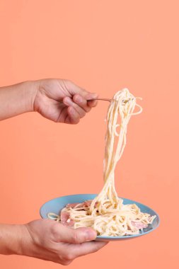 Turuncu arka planda izole edilmiş yeşil bir tabakta, çatallı kremalı spagetti yiyen. İtalyan yemeği, İtalyan mutfağı, erişte..