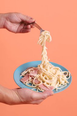 Turuncu arka planda izole edilmiş yeşil bir tabakta, çatallı kremalı spagetti yiyen. İtalyan yemeği, İtalyan mutfağı, erişte..