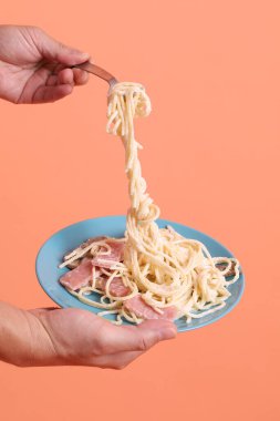 Turuncu arka planda izole edilmiş yeşil bir tabakta, çatallı kremalı spagetti yiyen. İtalyan yemeği, İtalyan mutfağı, erişte..