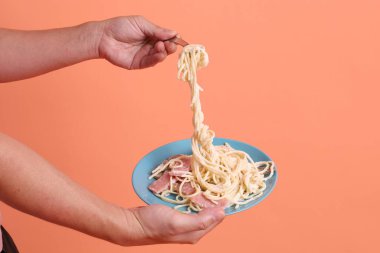 Turuncu arka planda izole edilmiş yeşil bir tabakta, çatallı kremalı spagetti yiyen. İtalyan yemeği, İtalyan mutfağı, erişte..