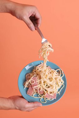 Turuncu arka planda izole edilmiş yeşil bir tabakta, çatallı kremalı spagetti yiyen. İtalyan yemeği, İtalyan mutfağı, erişte..