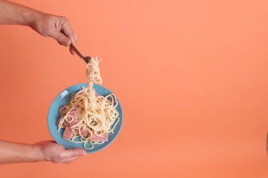 Turuncu arka planda izole edilmiş yeşil bir tabakta, çatallı kremalı spagetti yiyen. İtalyan yemeği, İtalyan mutfağı, erişte..