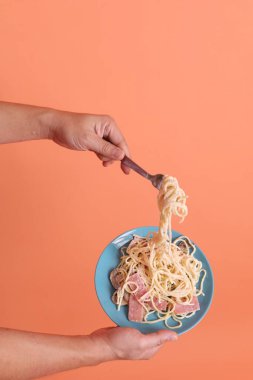 Turuncu arka planda izole edilmiş yeşil bir tabakta, çatallı kremalı spagetti yiyen. İtalyan yemeği, İtalyan mutfağı, erişte..