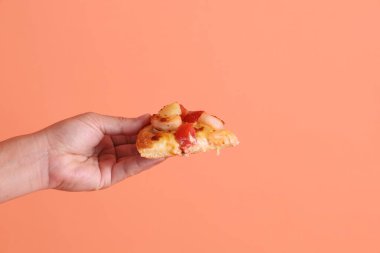 Elinde bir dilim Hawaii pizzası, üzerinde domates sosu, peynir, ananas ve domuz pastırması olan bir pizza, turuncu arka planda izole edilmiş beyaz bir tabakta. İtalyan yemeği, İtalyan mutfağı..