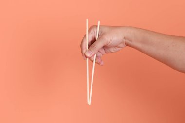 Elle tutulan ahşap çubuklar, turuncu bir arka planda izole edilmiş. Yemek ve yemek, Asya ve Çin kültürü, stüdyo çekimi..