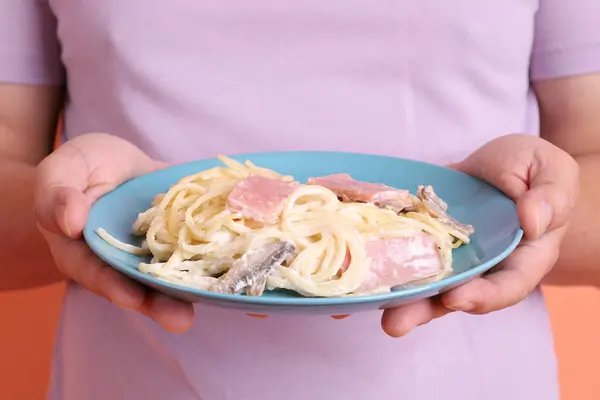 Elle tutulan kremalı spagetti carbonara yeşil bir tabakta, turuncu bir arka planda izole edilmiş. İtalyan yemeği, İtalyan mutfağı, erişte..