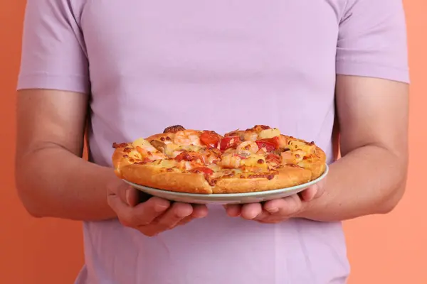 Elinde koca bir Hawaii pizzası, üzerinde domates sosu, peynir, ananas ve domuz pastırması olan bir pizza, turuncu bir arka planda izole edilmiş, beyaz bir tabakta. İtalyan yemeği, İtalyan mutfağı..