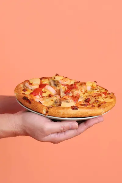 Elinde koca bir Hawaii pizzası, üzerinde domates sosu, peynir, ananas ve domuz pastırması olan bir pizza, turuncu bir arka planda izole edilmiş, beyaz bir tabakta. İtalyan yemeği, İtalyan mutfağı..
