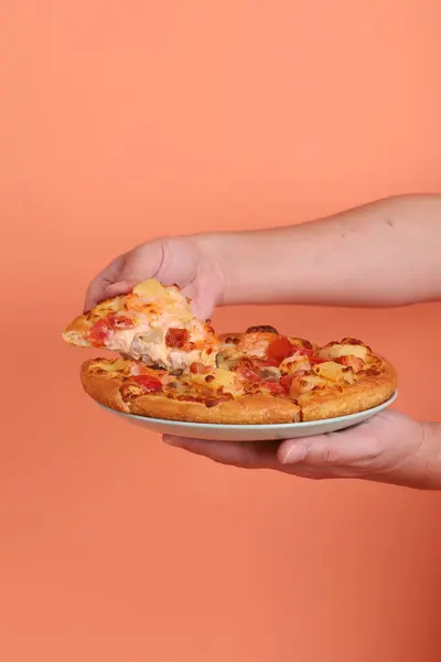 Elinde koca bir Hawaii pizzası, üzerinde domates sosu, peynir, ananas ve domuz pastırması olan bir pizza, turuncu bir arka planda izole edilmiş, beyaz bir tabakta. İtalyan yemeği, İtalyan mutfağı..