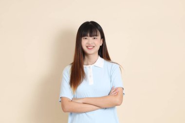 Açık mavi polo tişörtlü, beyaz şortlu, kolları çapraz duran ve bej arka planda güvenle gülümseyen genç bir Asyalı kadının orta boy fotoğrafı. Üniversite öğrencisi, Gen Z