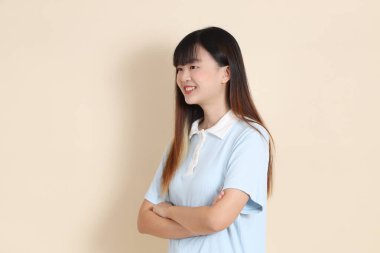 Açık mavi polo tişörtlü, beyaz şortlu, kolları çapraz duran ve bej arka planda güvenle gülümseyen genç bir Asyalı kadının orta boy fotoğrafı. Üniversite öğrencisi, Gen Z