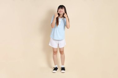 Açık mavi polo tişörtlü, beyaz şortlu, kolları çapraz duran ve bej arka planda güvenle gülümseyen genç bir Asyalı kadının vücut portresi. Üniversite öğrencisi, Gen Z