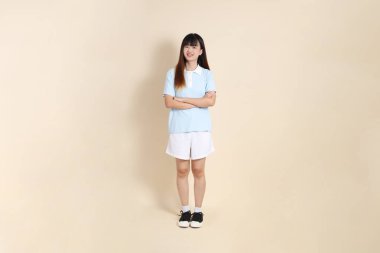 Açık mavi polo tişörtlü, beyaz şortlu, kolları çapraz duran ve bej arka planda güvenle gülümseyen genç bir Asyalı kadının vücut portresi. Üniversite öğrencisi, Gen Z