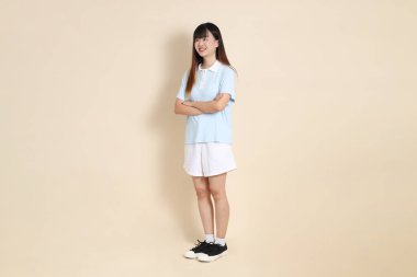 Açık mavi polo tişörtlü, beyaz şortlu, kolları çapraz duran ve bej arka planda güvenle gülümseyen genç bir Asyalı kadının vücut portresi. Üniversite öğrencisi, Gen Z