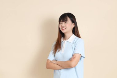 Açık mavi polo tişörtlü, beyaz şortlu, kolları çapraz duran ve bej arka planda güvenle gülümseyen genç bir Asyalı kadının orta boy fotoğrafı. Üniversite öğrencisi, Gen Z
