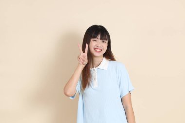 Orta boy Asyalı bir kadının açık mavi polo tişörtü, beyaz şort, barış işareti, V işareti, iki parmağı bej arka planda. Üniversite öğrencisi, Gen Z