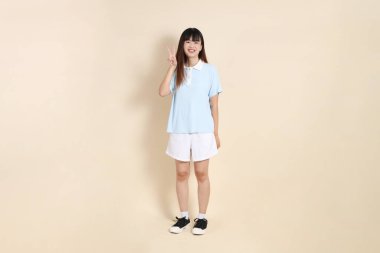 Açık mavi polo tişört giyen genç bir Asyalı kadının tüm vücut portresi, ve beyaz şort, barış işareti, V işareti, düz bir bej arka planda iki parmak. Üniversite öğrencisi, Gen Z