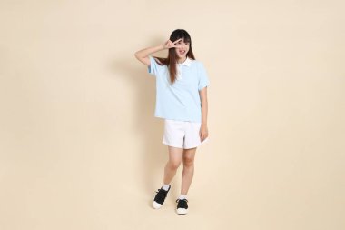 Açık mavi polo tişört giyen genç bir Asyalı kadının tüm vücut portresi, ve beyaz şort, barış işareti, V işareti, düz bir bej arka planda iki parmak. Üniversite öğrencisi, Gen Z
