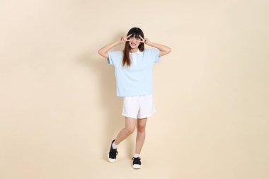 Açık mavi polo tişört giyen genç bir Asyalı kadının tüm vücut portresi, ve beyaz şort, barış işareti, V işareti, düz bir bej arka planda iki parmak. Üniversite öğrencisi, Gen Z